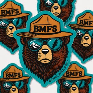 May include: A die-cut sticker featuring a brown bear wearing a tan ranger hat with the text BMFS printed on the front. The bear wears teal-rimmed sunglasses reflecting a mountain landscape and a lightning bolt. The text @COSMIC_RIPPLE_EFFECT is visible.