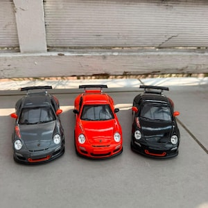 Könnte beinhalten: Drei Modellautos im Sportwagen-Design in einer Reihe. Das linke Auto ist metallic-grau, das mittlere leuchtend orange und das rechte schwarz. Jedes Fahrzeug verfügt über einen Heckspoiler, orangefarbene Seitenspiegel und orange Frontakzente.