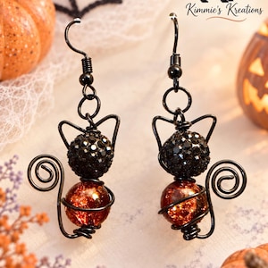 May include: A pair of dangle earrings featuring a cat-shaped wire frame in black metal. Each earring includes a black faceted bead for the head and an orange crackle glass bead for the body, with a coiled wire tail and pointed ears.