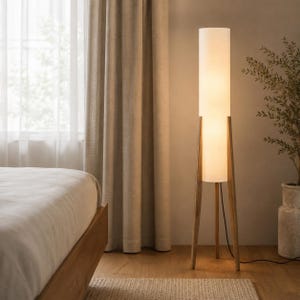 May include: A tall floor lamp featuring a cylindrical white fabric shade supported by three slender wooden legs. The lamp emits a soft, warm glow and stands on a wooden base, with a thin black power cord extending from the bottom of the structure.