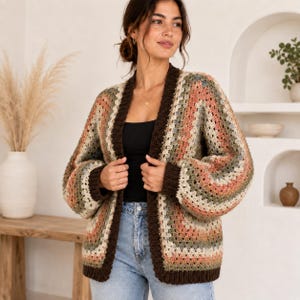 May include: A long-sleeve crochet cardigan featuring a multi-colored granny square pattern in shades of muted peach, sage green, cream, and brown. The garment has a solid dark brown ribbed trim along the open front, cuffs, and bottom hem.