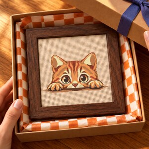 Puede incluir: Un bordado de punto de cruz cuadrado que muestra un gato atigrado naranja asomándose por un borde, enmarcado en madera de color marrón oscuro. La obra se presenta dentro de una caja de regalo de cartón forrada con papel de seda a cuadros naranja y blanco.