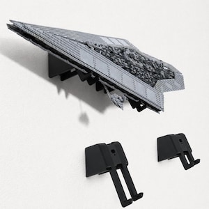 May include: A grey plastic building block model of a starship mounted on a black wall bracket, with two additional black wall-mounted brackets positioned below it. The model features a flat, triangular design with intricate surface detailing.
