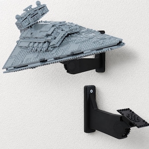 May include: A grey plastic model of a Star Destroyer spaceship mounted on a black wall bracket, with a second identical black wall bracket positioned below it. The brackets feature a flat mounting plate and a gear-like adjustable joint mechanism.