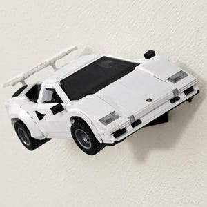 May include: A white toy sports car model constructed from building blocks, featuring a rear spoiler, black windshield, and black tires. The vehicle has a low-profile design with angular body panels and a small emblem on the front hood.