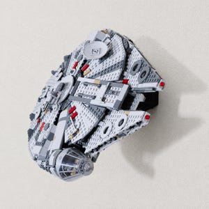 May include: A detailed model of the Millennium Falcon spaceship constructed from gray, white, and dark gray plastic building bricks. The structure features a circular hull, a cockpit with a transparent dome, and small red accents on the exterior surface.