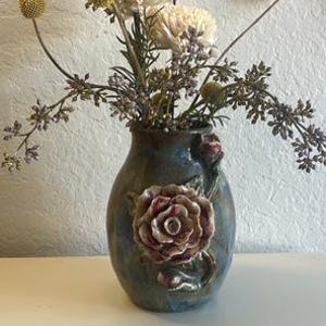 May include: A ceramic vase with a mottled blue and green glaze featuring a three-dimensional sculpted rose on the front. The vase is filled with an arrangement of dried cream-colored flowers, yellow billy ball stems, and small purple sprigs.