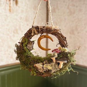 Puede incluir: Un adorno de corona de ramitas con una pequeña figura de búho marrón, una luna creciente amarilla y dos setas con sombreros rojos y blancos. La corona está decorada con musgo verde y trozos de corteza de árbol, suspendida por un cordel.