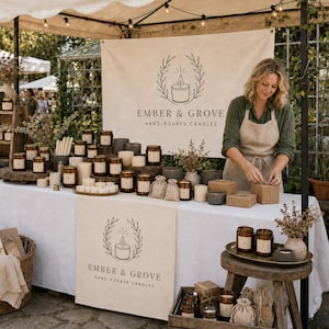 May include: A market stall display featuring Ember & Grove hand-poured candles in various glass jars and small fabric pouches. The setup includes a white tablecloth, wooden shelving, a wicker basket, and a banner with the brand logo and text.
