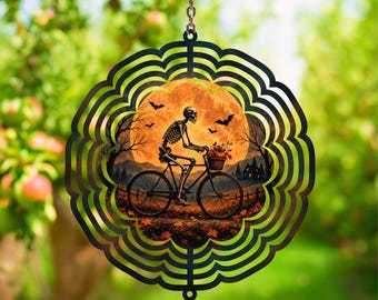 May include: A circular metal wind spinner featuring a black scalloped frame and a central graphic of a skeleton riding a bicycle. The scene depicts a large orange full moon, flying bats, bare trees, and fallen autumn leaves on the ground.