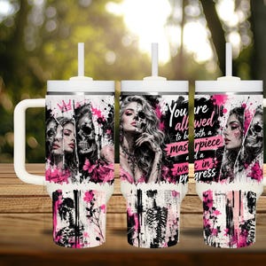 May include: A white tumbler with a handle and straw featuring a graphic print of a woman with a skull face, pink paint splatters, and floral accents. The text reads, You are allowed to be both a masterpiece and a work in progress.