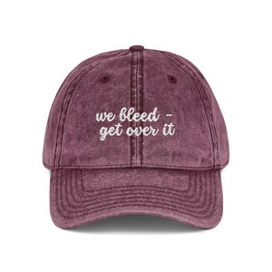 Könnte beinhalten: Eine kastanienbraune Baseballkappe mit verwaschener, abgenutzter Textur. Auf der Vorderseite befindet sich ein weißer Stickschriftzug mit dem Text "we bleed - get over it" in Schreibschrift. Die Kappe hat eine strukturierte Krone und einen Schirm.