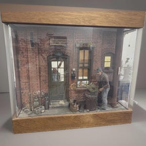 May include: A miniature diorama of a brick building exterior labeled Leroy & Bertha's Bar and Grill, enclosed in a wooden display case. The scene features a person standing near a trash can, a door with a window, and a window with yellow light.