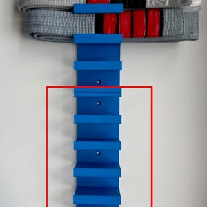 May include: A blue vertical wall-mounted rack with multiple horizontal slots designed for holding martial arts belts. The rack features a series of evenly spaced shelves and pre-drilled holes for mounting, with three folded belts resting on the top.