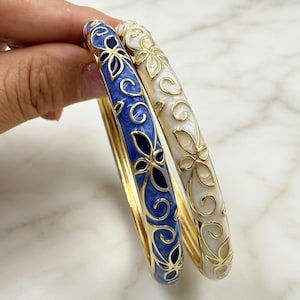 May include: Two bangle bracelets featuring gold-tone metal filigree overlays. One bracelet has a deep blue enamel base, while the other has a cream-coloured enamel base. Both bangles are decorated with repeating floral and swirling patterns.