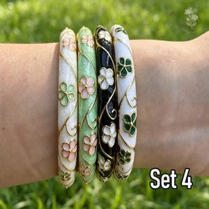 May include: A set of four bangle bracelets featuring enamel finishes in white, light green, black, and white. Each bracelet is decorated with gold-toned floral patterns and swirling vine motifs. The bangles are arranged side by side in a row.