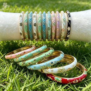 Peut inclure: Une collection de bracelets joncs en métal ornés de motifs floraux en émail de différentes couleurs, notamment blanc, bleu clair, vert menthe, rose, lavande et rouge. Les bracelets sont présentés sur un support cylindrique blanc et sur l'herbe.