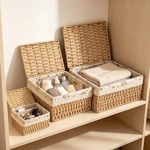 May include: Three rectangular woven wicker storage baskets with hinged lids and fabric liners. The liners feature a floral pattern and white lace trim. The baskets contain various personal items, including bottles, folded fabric, and small accessories.