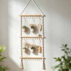 Peut inclure: Une décoration murale suspendue composée de trois tiges en bois clair reliées par un cordon en macramé crème. L'ensemble comporte des crochets métalliques dorés, des petites plantes succulentes, des fleurs séchées, une feuille et un oiseau.