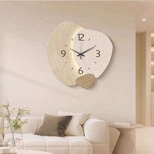 Peut inclure: Une horloge murale composée de trois panneaux organiques superposés en forme de galets dans des tons beige et crème. Le cadran affiche des chiffres noirs aux positions 3, 6, 9 et 12 heures avec des aiguilles fines et un éclairage intégré.