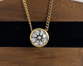 May include: A gold necklace featuring a round, clear, faceted stone set in a smooth, circular bezel setting. The pendant is suspended from a delicate gold curb chain, showcasing a brilliant cut with a star-like reflection pattern at the center.