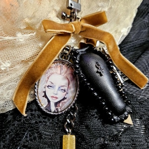 May include: A keyring featuring a gold velvet ribbon bow, a silver-tone lobster clasp, an oval pendant with a portrait of a pale figure with fangs, a small black coffin charm with a cross, and a rectangular yellow bead on a dark metal chain.