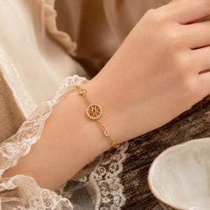Puede incluir: Una pulsera de cadena en tono dorado con un dije circular central diseñado como una brújula o un timón de barco, incrustado con pequeñas piedras claras. La delicada cadena está acentuada con piedras claras adicionales a lo largo.