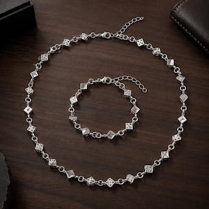 Puede incluir: Un conjunto de collar y pulsera en tono plateado que presenta una serie de pequeños dijes en forma de dados cuadrados unidos por anillos metálicos circulares. Ambas piezas incluyen un cierre de mosquetón y una cadena de extensión ajustable.