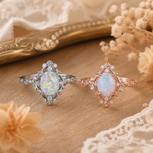 Puede incluir: Dos anillos con ópalos de corte ovalado que muestran destellos iridiscentes. Un anillo es de metal plateado y el otro de metal en tono oro rosa. Ambos engastes están adornados con pequeñas piedras transparentes de corte redondo en un marco.