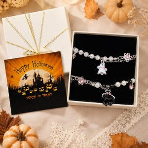 Peut inclure: Deux bracelets argentés ornés de perles blanches et de cristaux roses dans une boîte cadeau blanche. Un bracelet porte une breloque fantôme blanc, l'autre une breloque chapeau de sorcière noir. Une carte carrée avec une scène d'Halloween est incluse.