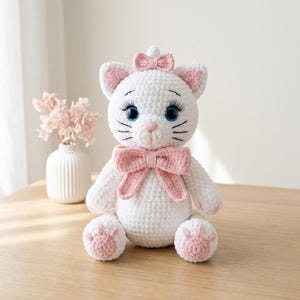 Puede incluir: Un peluche de gato tejido a ganchillo en color blanco con grandes ojos azules, largas pestañas negras, una nariz rosa bordada y orejas de color rosa en el interior. El gato lleva un lazo rosa en la cabeza y otro más grande en el cuello.
