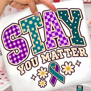 May include: A white t-shirt featuring the text STAY YOU MATTER in large, multi-patterned letters. The S and A have a purple gingham pattern, the T and Y have a teal floral print, and the T has a purple checkerboard pattern. Includes a teal and purple ribbon.
