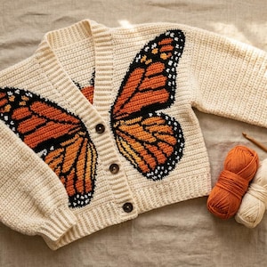 May include: A cream-colored crochet cardigan featuring a large orange, black, and white monarch butterfly wing pattern across the front panels. The garment has long sleeves, a ribbed V-neckline, and three dark brown buttons. Includes orange and cream yarn.