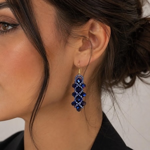 May include: A dangling earring featuring a vertical arrangement of deep blue faceted crystal beads and smaller metallic blue seed beads. The design consists of a repeating woven pattern suspended from a simple gold-toned metal hook wire.
