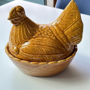 Peut inclure: Un plat à couvercle en céramique jaune moutarde brillant en forme de poule. Le couvercle présente une poule sculptée avec des détails de plumes, tandis que la base est conçue pour ressembler à un panier tressé. Finition lisse et brillante.