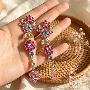 Può includere: Un paio di orecchini pendenti lunghi con motivo floreale, realizzati in metallo dorato e strass multicolori. Ogni orecchino presenta cristalli circolari rosa, viola e azzurri disposti a forma di fiore, con accenti a forma di foglia verde chiaro.