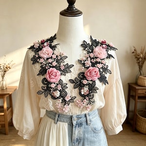 May include: A cream-colored blouse featuring black lace embroidery and three-dimensional pink fabric roses on the chest. The garment has long sleeves, a V-neckline, and is paired with light blue denim jeans. The floral embellishments are symmetrical.