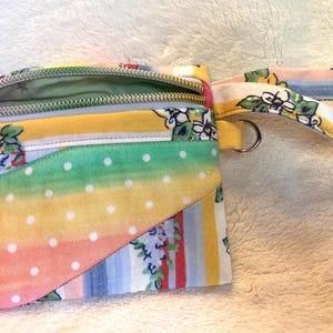 May include: A small fabric wristlet pouch featuring a multi-colored pattern with yellow, blue, and pink stripes, floral motifs, and a curved front pocket section decorated with white polka dots on a gradient background, including a matching wrist strap.