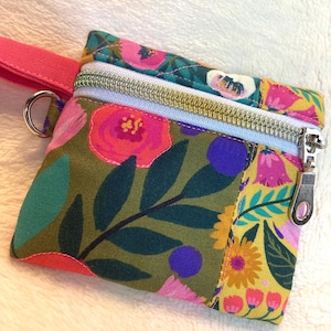 May include: A small fabric pouch featuring a vibrant floral pattern with pink, yellow, and green tones. The pouch includes a silver-toned metal zipper closure, a matching pink wrist strap attached to a silver metal ring, and a light blue zipper tape.