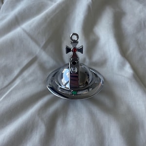 Può includere: Un ciondolo sferico color argento sormontato da una croce con una piccola pietra rossa al centro. Il corpo sferico è circondato da un anello e decorato con un cristallo trasparente e una pietra verde, con una finitura metallica lucida.