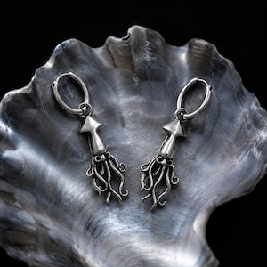 May include: A pair of silver-toned hoop earrings featuring dangling squid charms. Each charm displays a detailed mantle, head, and multiple flowing tentacles. The metal has a polished finish with darkened recessed areas to highlight the anatomical features.