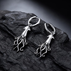 May include: A pair of silver-toned hoop earrings featuring dangling squid charms. Each charm is shaped like a squid with a smooth mantle and multiple curved tentacles extending downward. The earrings have a polished metallic finish and a hinged hoop closure.