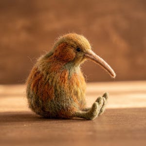 Può includere: Una statuetta di uccello kiwi in lana infeltrita con un corpo arrotondato e un becco lungo e sottile. L'uccello presenta una superficie testurizzata e soffice con un motivo a colori screziati di marrone terra, arancione ruggine e verde oliva.