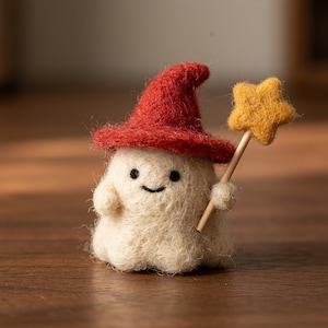 Puede incluir: Una pequeña figura de fieltro de aguja de un fantasma blanco con un sombrero puntiagudo rojo. El fantasma tiene dos ojos de cuentas negras y una sonrisa negra cosida. Sostiene una estrella de fieltro amarilla en un palo de madera.