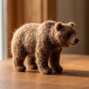 Peut inclure: Une figurine d'ours en laine feutrée à l'aiguille, composée de fibres de laine brunes texturées. L'ours est représenté en position debout, arborant un pelage dense et duveteux, de petites oreilles arrondies et un museau brun foncé.