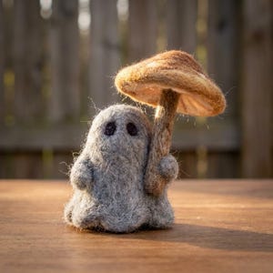 Peut inclure: Une figurine de fantôme gris en laine feutrée avec deux petits yeux noirs, tenant un objet en forme de champignon brun et beige. Le fantôme a un aspect duveteux et texturé, se tenant debout avec le chapeau du champignon au-dessus de sa tête.