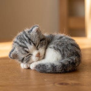 Peut inclure: Une sculpture en laine feutrée représentant un chaton endormi au pelage gris et blanc. Le chaton est recroquevillé en boule, les yeux fermés, avec un petit nez rose et des fibres de laine texturées et duveteuses sur tout le corps et les oreilles.
