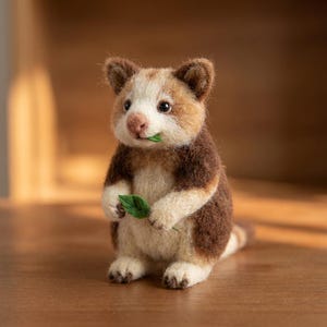 May include: A needle-felted animal figurine with a cream-coloured face, dark brown back, and light brown ears. The creature has black eyes, a pink nose, and holds a small green fabric leaf in its paws, with another leaf in its mouth.