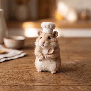 May include: A needle-felted hamster figurine with light brown and white fur, wearing a small white chef hat on its head. The hamster is holding a tiny wooden spoon in its front paws, featuring a pink nose, black eyes, and visible whiskers.