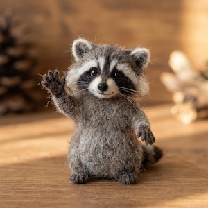 May include: A needle-felted raccoon figurine with a fuzzy grey and white coat, black eye patches, and a small black nose. The raccoon is standing upright with one front paw raised in a waving gesture, featuring detailed whiskers and dark paws.
